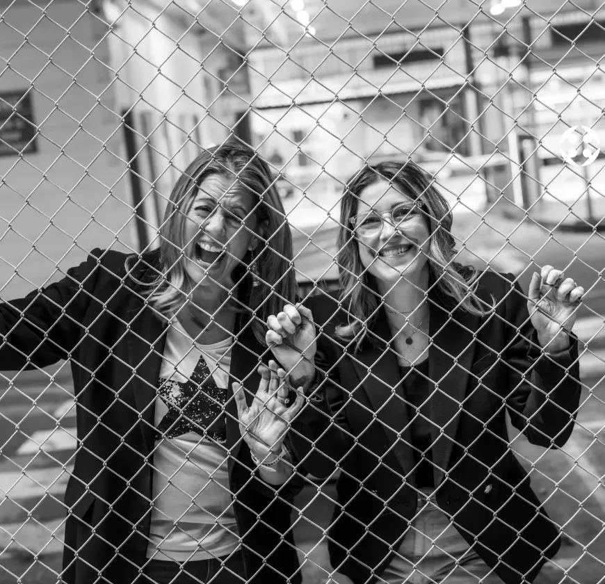 Emily Jaksch and Jessy Warn laughing behind a wire fence, in black and white.